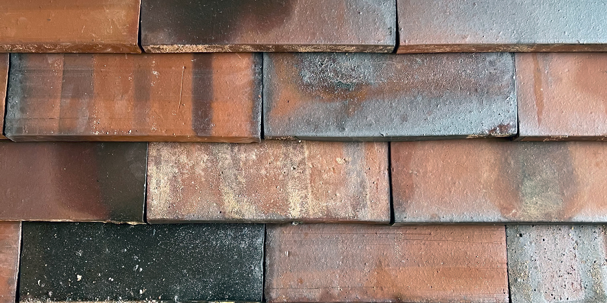 Musterwand mit 3 Reihen großfächiger Keramikschindeln welche leicht überlappend montiert sind. Die Keramikschindeln sind rot-brau-blaulich mit glänzenden Kohleaufbrennungen und das Ganze wirkt sehr lebendig